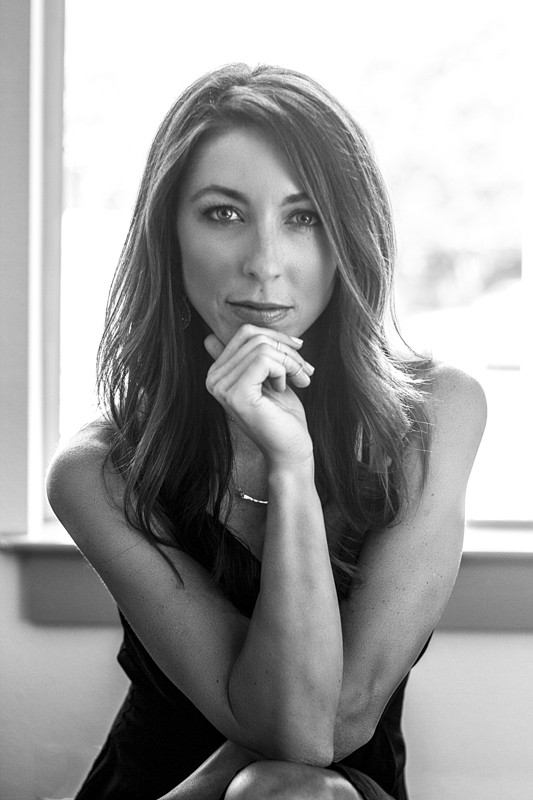 Black and white portrait of a woman with long brown hair resting her chin on her hand near a window