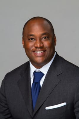 Smiling man in a dark suit and blue tie posing for a professional headshot against a gray background