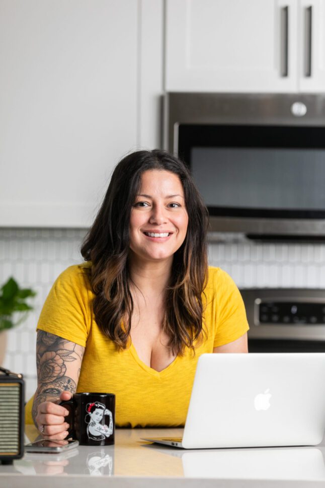 Woman in a yellow top smiling at a kitchen counter with a coffee mug and laptop