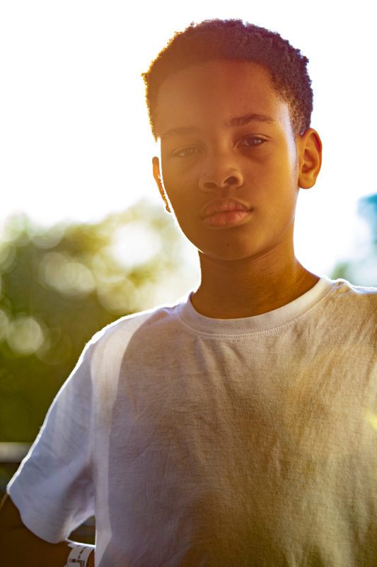 Young boy in a white t-shirt standing outdoors with warm backlit sunlight through the trees