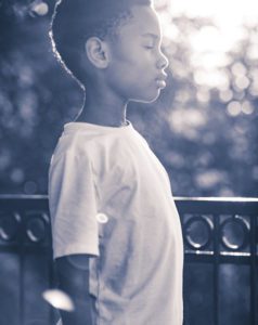 Black and white backlit profile portrait of a young boy standing outdoors with sunlight through the trees
