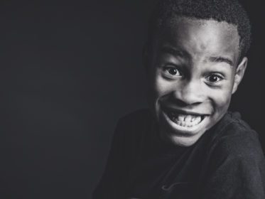 Black and white portrait of a young boy grinning widely and looking down at the camera against a dark background