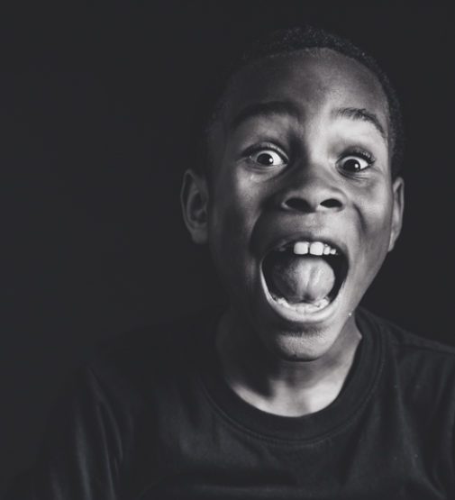 Black and white portrait of a young boy making a silly, exaggerated shouting face against a dark background