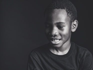 Black and white close-up candid portrait of a young boy smiling and looking down against a dark background