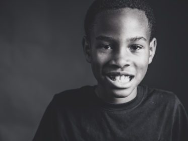 Black and white close-up portrait of a young boy smiling with his teeth showing against a dark background