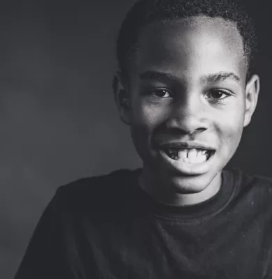 Black and white close-up portrait of a young boy smiling with his teeth showing against a dark background