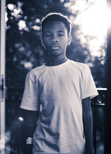 Black and white portrait of a young boy in a white t-shirt standing on a balcony with sunlight behind him