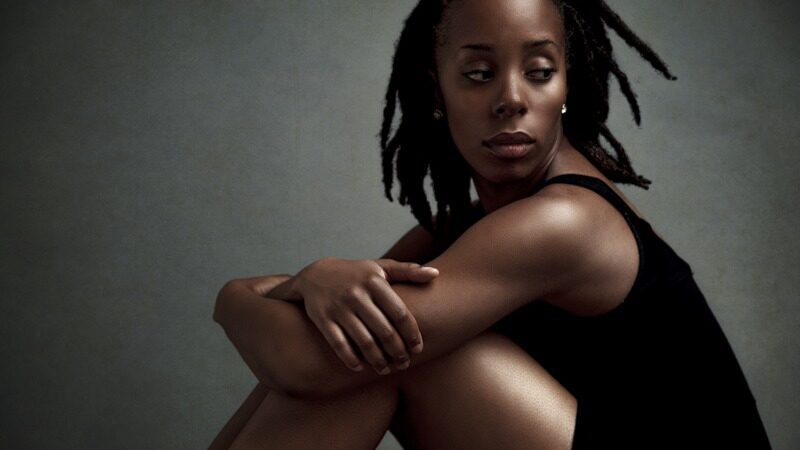 Woman with dreadlocks seated and hugging her knees in a soft dramatic studio portrait, representing self esteem and self love