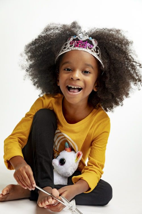 Laughing young girl with a large curly afro wearing a tiara and holding a unicorn stuffed toy