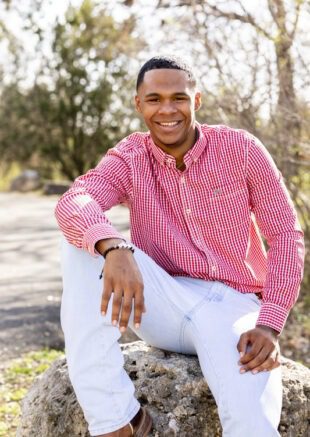 Smiling young man in a red gingham shirt and white jeans seated on a rock outdoors for his senior portrait
