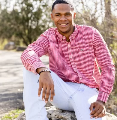 Smiling young man in a red gingham shirt and white jeans seated on a rock outdoors for his senior portrait