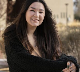 Smiling young woman with long dark hair wearing a black cardigan seated outdoors for her senior portrait