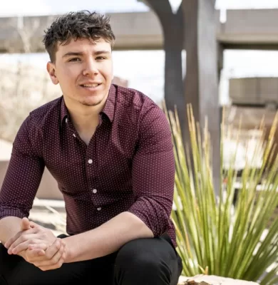 Smiling young man in a maroon patterned shirt seated outdoors with clasped hands for his graduation portrait