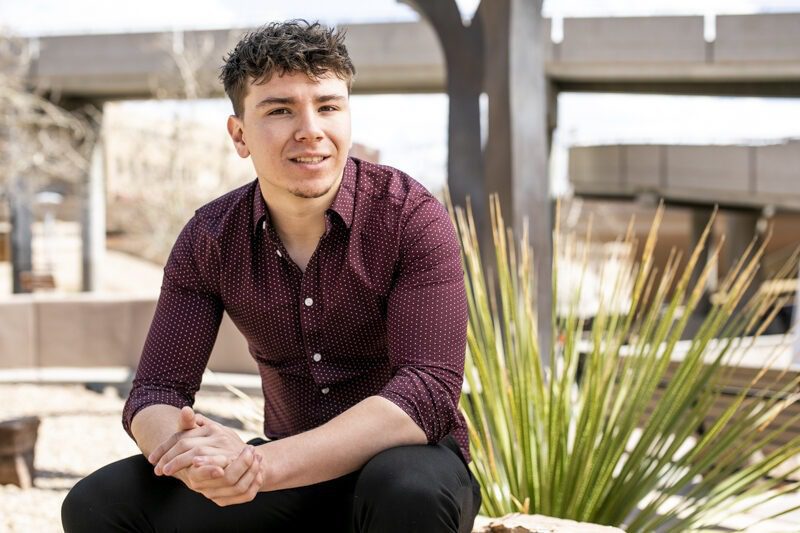 Smiling young man in a maroon patterned shirt seated outdoors with clasped hands for his graduation portrait