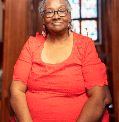 Elderly woman in a red top smiling for a portrait in front of stained glass windows