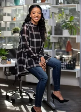 Smiling woman with curly hair wearing a plaid top and jeans seated on a stool in a plant-filled office