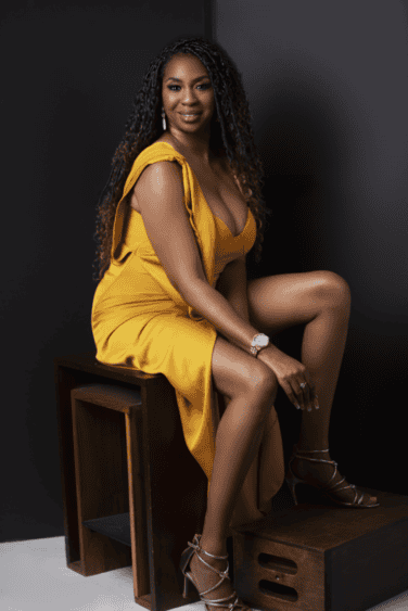 Smiling woman with curly hair wearing a yellow dress seated on a wooden stool in a dramatic studio portrait