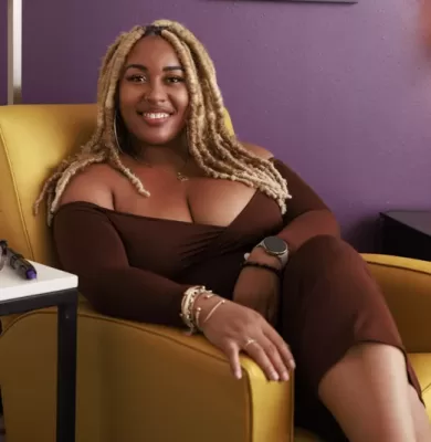 Smiling woman with blonde braids wearing a brown off-shoulder dress seated in a yellow armchair against a purple wall