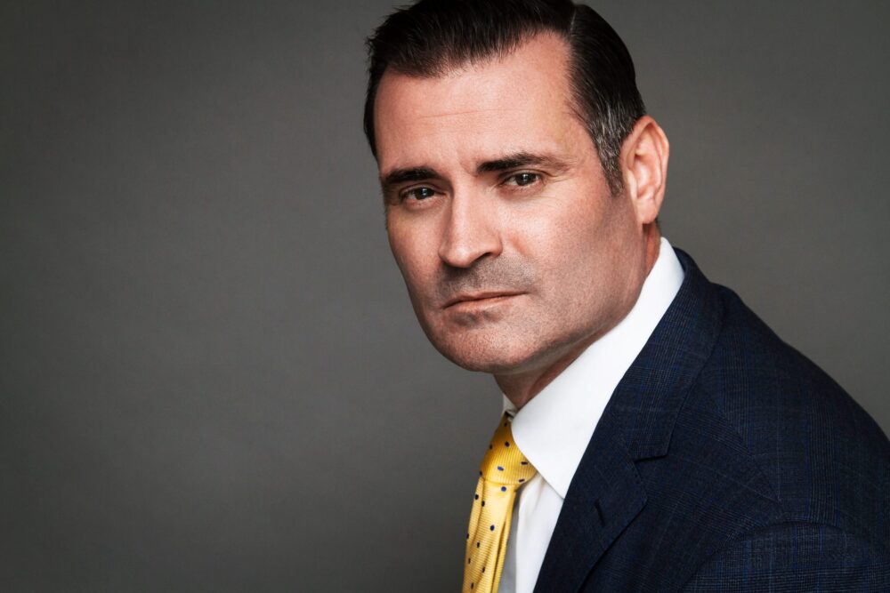 Man in a navy suit and yellow tie posing for a dramatic studio portrait against a gray background