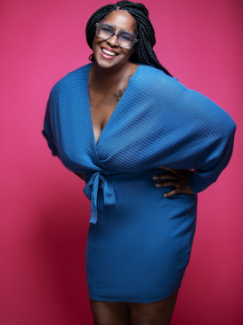 Laughing woman with braided hair wearing a blue textured wrap dress against a bright pink background
