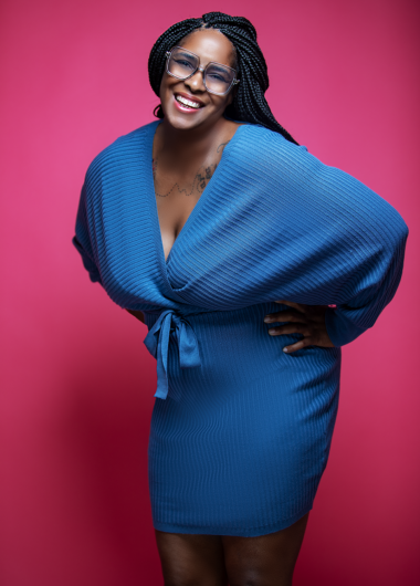 Laughing woman with braided hair wearing a blue textured wrap dress against a bright pink background