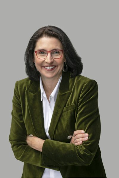 Smiling woman with red glasses wearing an olive velvet blazer, arms crossed, in a professional headshot