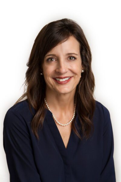 Professional headshot of a smiling woman with dark hair wearing a navy blouse and pearl necklace against a white background