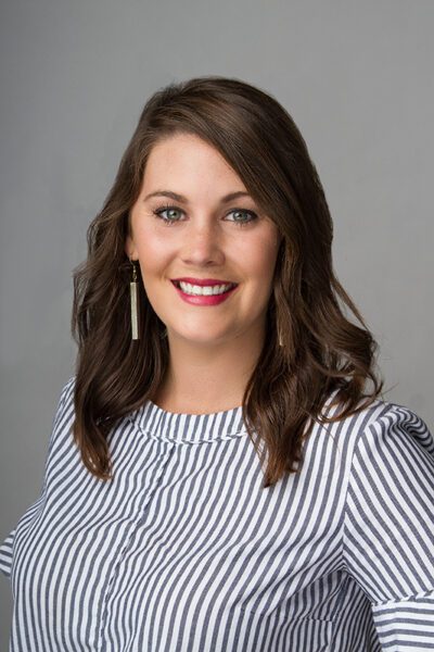 Professional headshot of a smiling woman with wavy brown hair wearing a striped blouse against a gray background