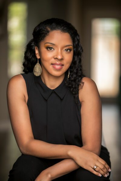 Smiling woman with curly hair wearing a black sleeveless top and drop earrings, seated for a professional real estate headshot