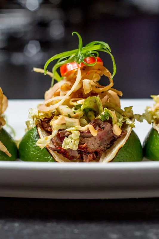 Close-up of a brisket taco topped with slaw and fried onions, garnished with lime wedges
